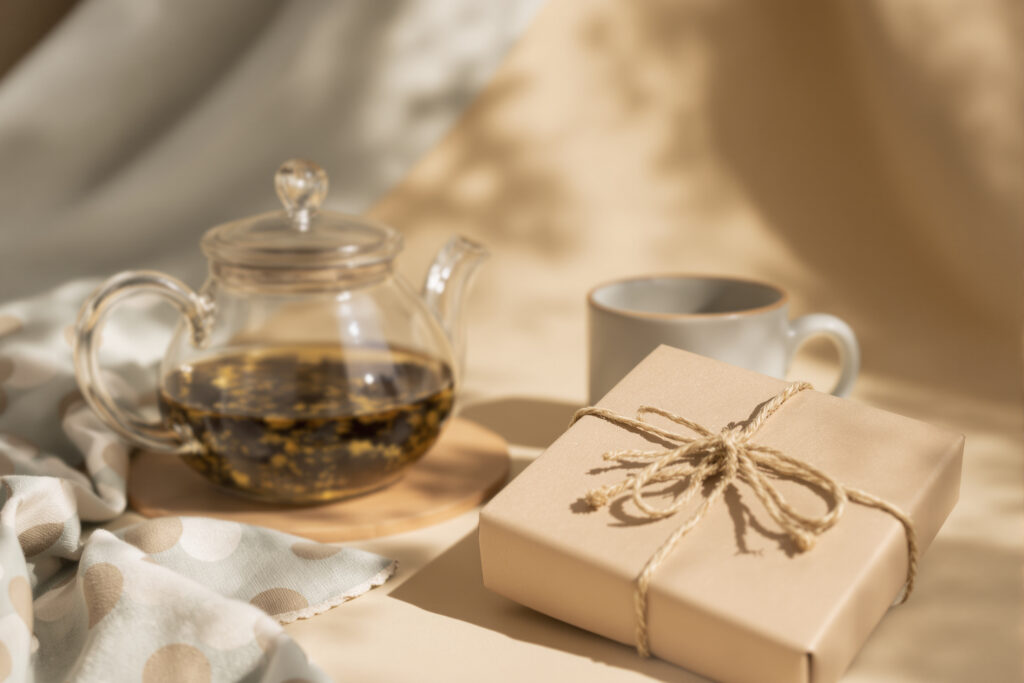 Tetera de cristal con té en hebras junto a una taza de gres y una vela envueltas para regalo